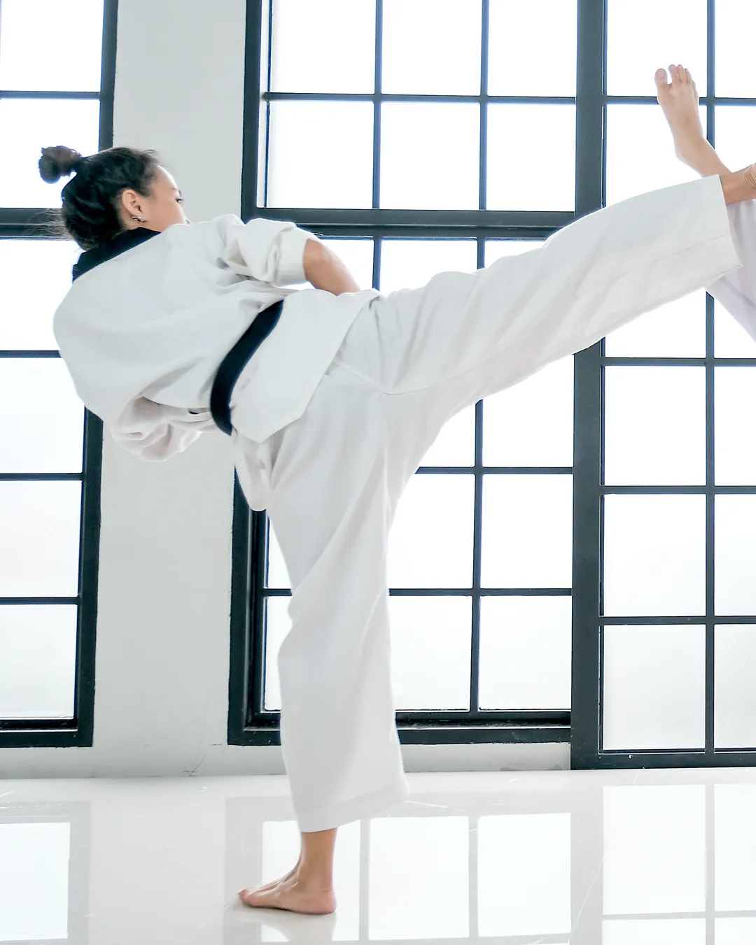 An adult student in a white Taekwondo uniform holds a high kick in a training room