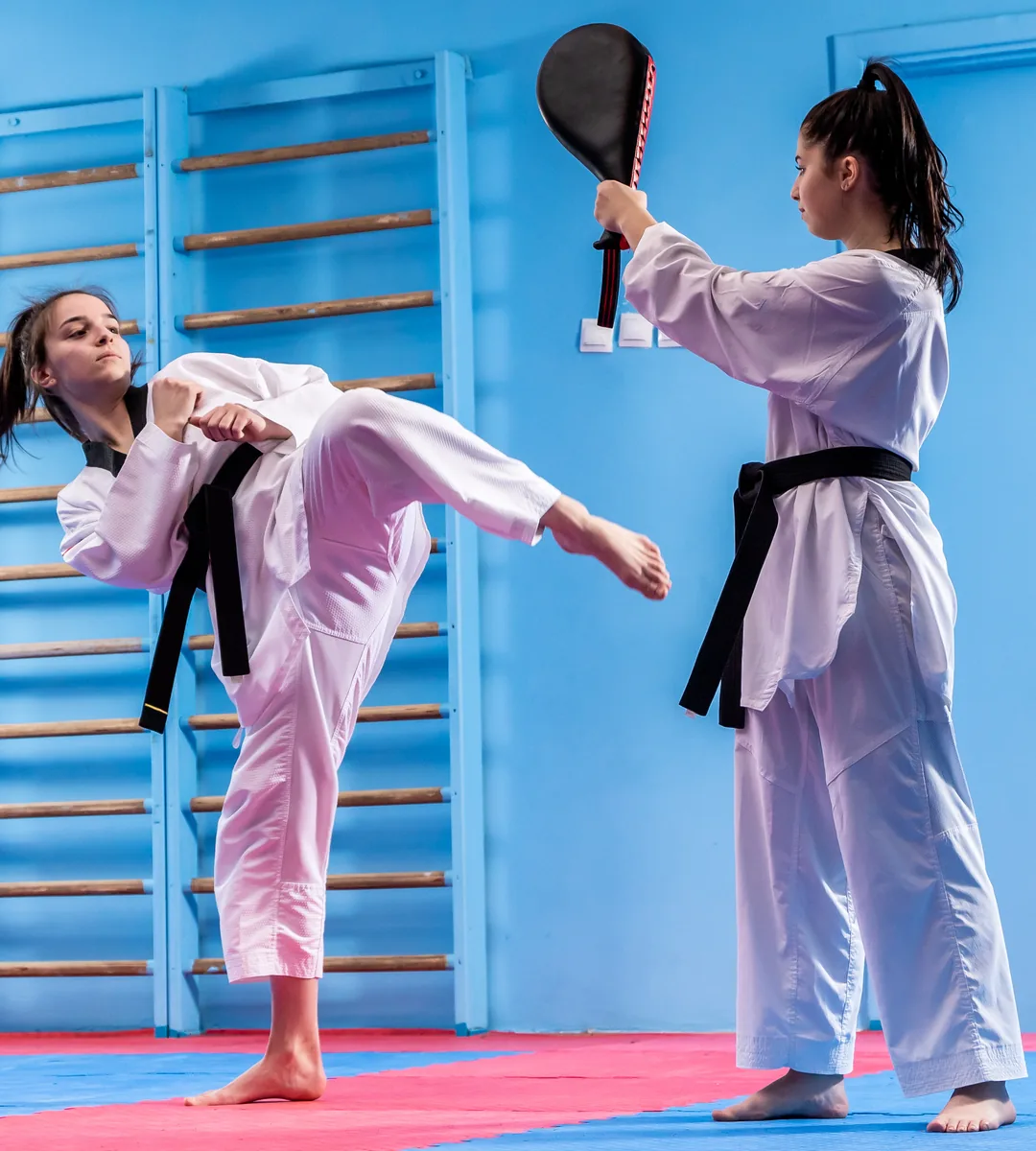 A woman in a Taekwondo uniform throws a high roundhouse kick in a wood-floored gym